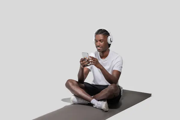 白いTシャツとヘッドフォンを着た若いアフリカ系アメリカ人男性がマットの上に横たわり 楽しい表情でスマートフォンを見つめている — ストック写真