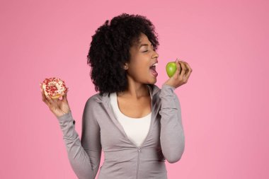 An african american lady is making a choice between a sugary donut and a healthy apple, showing a concept of diet decisions isolated on a pink backdrop clipart