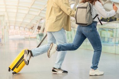 Sıradan giyinmiş iki kişi, biri sarı bavulu çekiyor, hava terminalinden geçiyor..