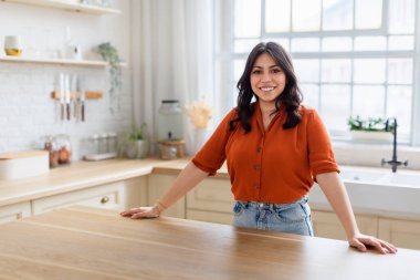 Parlak, çağdaş bir mutfakta kendine güvenen uzun saçlı neşeli bir genç kadın.