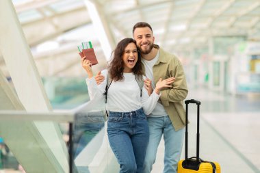 Havalimanında uçuş beklerken eğlenen heyecanlı genç adam ve kadın, bavulları terminalde bekleyen, pasaportlarını elinde tutan ve kameraya bakan mutlu çift.
