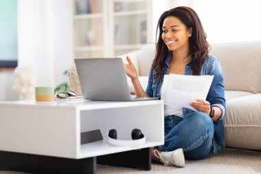 Afro-Amerikalı bir kadın evinde hem bilgisayarıyla hem de belgeleriyle meşgul, potansiyel bir çalışma ya da çalışma senaryosuyla.