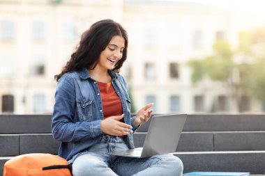 Neşeli zoomçu Hintli kadın laptopunu açık havada kullanıyor. Bağlantı ve kampüs hayatını simgeliyor. Boşluğu kopyala.