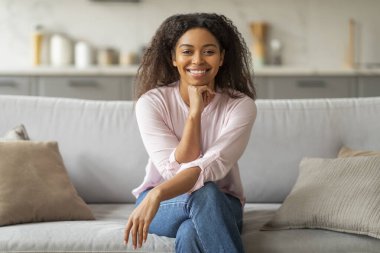 Güzel gülüşü olan Afro-Amerikalı bir bayan parlak, çağdaş bir evdeki kanepede zarif bir şekilde oturuyor.
