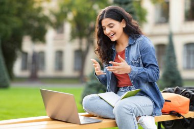 Exuding a sense of joy, an Indian lady laughs while looking at her laptop on a campus bench, highlighting the educational engagement and lively spirit of an eastern zoomer in an academic setting clipart