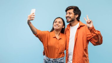 Turuncu gömlekli neşeli bir Hintli erkek ve kadın akıllı telefonlarıyla selfie çekiyorlar, gülümsüyorlar ve barış işareti ile poz veriyorlar.