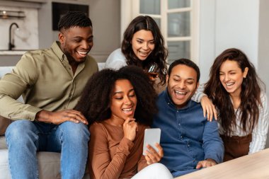 Bir grup çok ırklı, genç arkadaş birlikte gülüyor, akıllı telefona bakıyor, evinde rahat vakit geçiriyor.