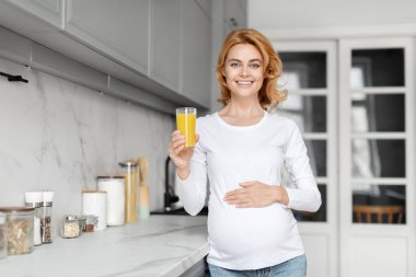A radiant european pregnant lady enjoys orange juice in a stylish kitchen, showcasing the importance of fruit in prenatal nutrition clipart
