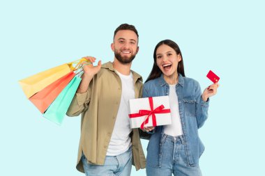 Renkli alışveriş çantaları taşıyan neşeli genç bir çift ve açık mavi arka planda bir kredi kartı sunan bir hediye kutusu.