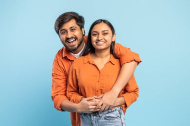 A smiling Indian man and woman warmly embracing each other, both wearing vibrant orange shirts against a blue background clipart