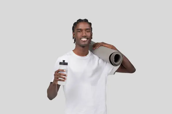 彼の腕の下にヨガマットと灰色の背景に対して彼の手の水瓶と一緒に立っている陽気な若い黒人男性 — ストック写真