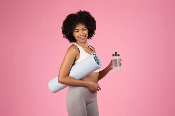 An athletic African American lady carrying a yoga mat and a water bottle ready for a workout session, isolated on a pink background