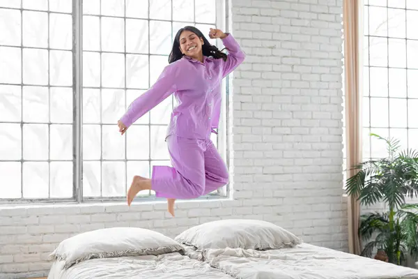 An exuberant middle eastern woman in purple pajamas captured mid-jump on her bed, portraying happiness and energy