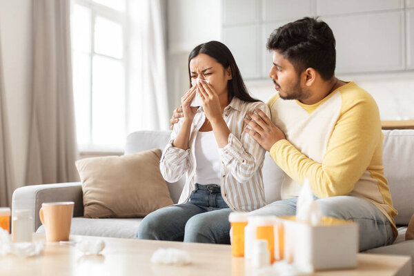 An Indian couple at home, the man comforting his sneezing spouse, depicting millennials managing health together