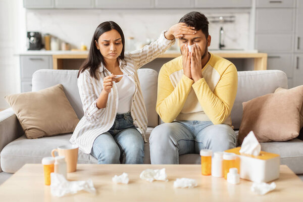 A concerned Indian woman supports her unwell spouse at home, representing eastern millennials coping with sickness