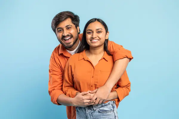 A smiling Indian man and woman warmly embracing each other, both wearing vibrant orange shirts against a blue background