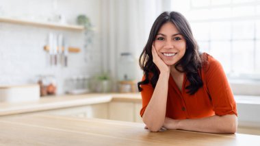 Uzun saçlı neşeli genç bir ortadoğulu kadın aydınlık modern bir mutfakta ışıl ışıl gülümsüyor, pozitiflik ve rahatlık saçıyor.