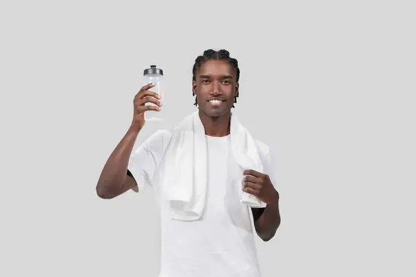 アフリカ系アメリカ人男性は笑顔でスポーツウォーターボトルを持って肩にタオルを引っ張り 平らな背景に隔離された — ストック写真