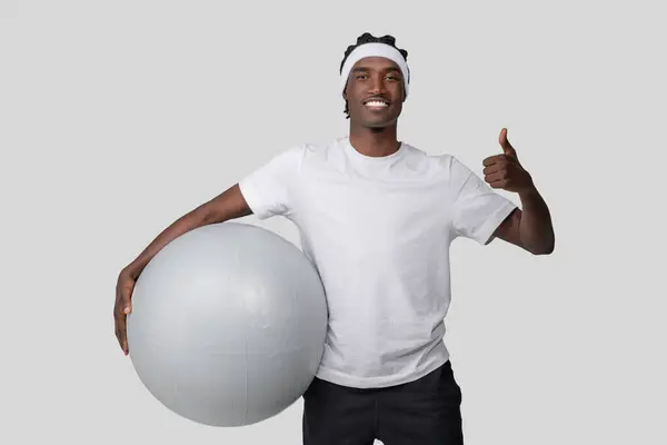 スポーツウェアの陽気なアフリカ系アメリカ人男性は 灰色の背景に親指を与えながら 自信を持って大きな運動ボールを保持しています — ストック写真