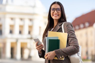 Mutlu genç kız öğrenci, elinde kitaplarla üniversitenin dışında akıllı telefonuyla iletişim kuruyor.