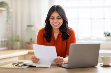 Turuncu bluzlu gülümseyen ortadoğulu kadın evinde dizüstü bilgisayarının yanında bir belgeyi zevkle inceliyor.