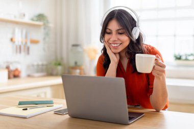 Young middle eastern woman with headphones using laptop and enjoying a cup of coffee, embodies multitasking at home clipart