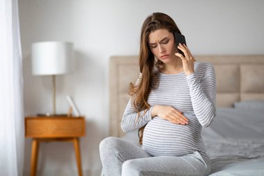 Hasta hamile kadın doktorla telefonda konuşurken stresli görünüyor. Yatakta karnını tutarak oturuyor.