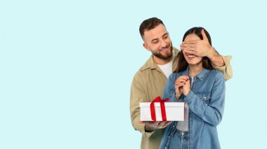 Sakallı bir adam, bir kadının gözlerini kapatırken ve elinde kırmızı kurdeleli bir hediye kutusu tutarken görülüyor. Mavi arka planda sürpriz bir hediye olması muhtemel.