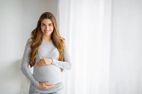 stock image A happy and radiant pregnant woman with long hair in cozy clothing is standing by a white curtain, cradling her belly