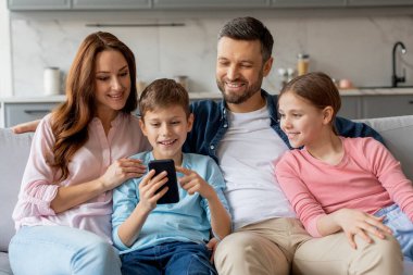 Dört kişilik neşeli bir aile kanepede sarılıyor ve akıllı bir telefona bakıyor, bir anlık bağlanma ve teknolojik etkileşim sergiliyor.