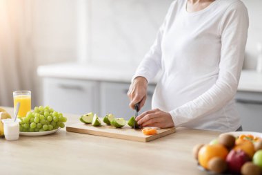 Beyaz gömlekli hamile bir Avrupalı kadın modern mutfağında besleyici bir meyve salatası hazırlıyor.