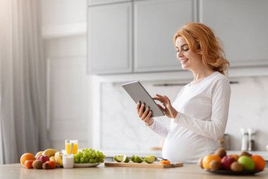 Avrupa özellikleri olan hamile bir kadın çeşitli besleyici mutfak malzemeleriyle çevrili tablete bakıyor.