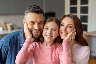 A smiling family with a daughter in the middle hugging her parents in a modern kitchen setting. They seem connected and joyful clipart