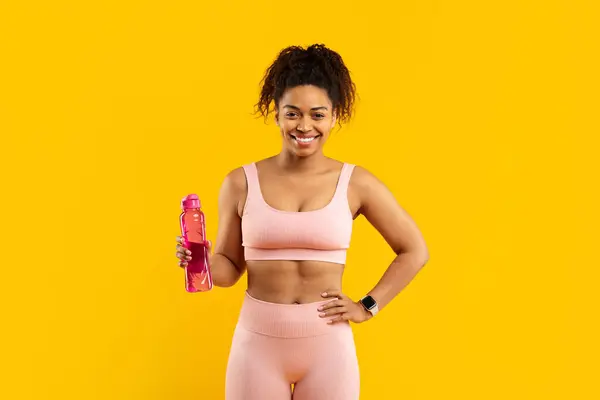 ピンクのスポーツウェアの陽気な若いアフリカ系アメリカ人女性は 明るい黄色の背景に再利用可能なウォーターボトルを保持し トレーニングの準備ができています — ストック写真