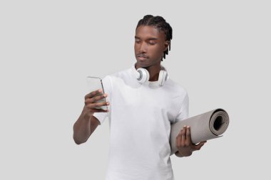 A young African American man holds a smartphone and carries a yoga mat, suggesting a lifestyle that combines technology and fitness clipart