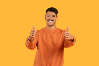 A millennial man with a moustache and a funny expression gives two thumbs up in a radiant orange sweater, isolated against an orange backdrop clipart