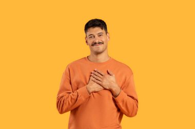 Turuncu gömlekli neşeli bir gencin samimi bir portresi, elleri yüreğinde, canlı sarı bir arkaplanda.