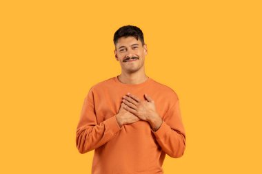 A candid portrait of a cheerful young man in an orange shirt, hands on heart, against a vibrant yellow background clipart