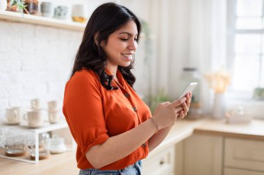 Gülümseyen Orta Doğulu kadın, güneşli bir mutfakta akıllı telefonuyla mesajlaşmaktan hoşlanıyor, bağlantı ve neşe tasvir ediyor.
