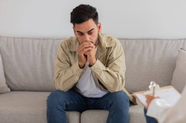 Genç bir adam terapi seansında düşünceli ve endişeli görünüyor. Depresyon, anksiyete ve travma sonrası stres bozukluğu çekiyor.