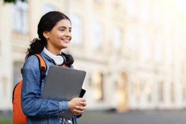 Kulaklık ve dizüstü bilgisayarı olan Hintli bir kadının kampüs atmosferinde eğitim ve teknolojiyi yakınlaştırması.