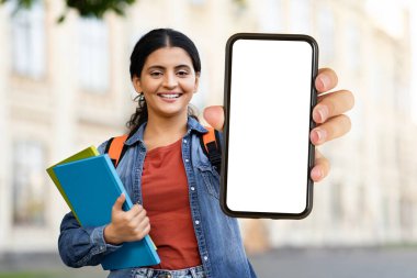 Sırt çantası ve kitapları olan neşeli genç bir kadın açık havada duran beyaz beyaz ekranlı bir cep telefonu sergiliyor.