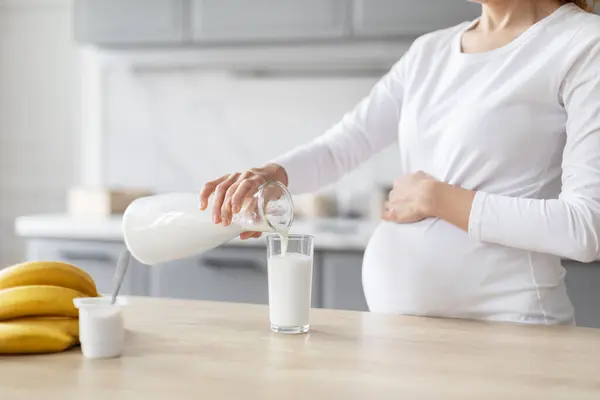 一位欧洲裔孕妇被描绘在厨房里 当她把牛奶倒入杯子里 四周都是新鲜的水果时 她把注意力集中在营养上 — 图库照片