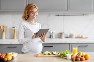 Avrupalı hamile bir kadın modern bir mutfakta duruyor ve etrafını taze besinlerle çevrili bir tablete bakıyor.