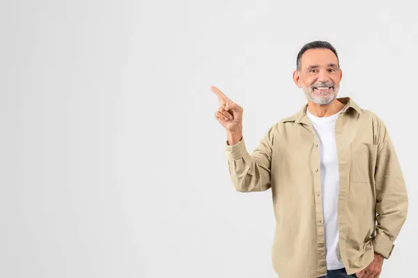 stock image A bearded senior man confidently points upwards, dressed casually in a beige shirt and white t-shirt, with an isolated white background, copy space
