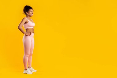 A toned african american woman poses in fitness wear with hands on her hips, isolated on a yellow background, copy space clipart