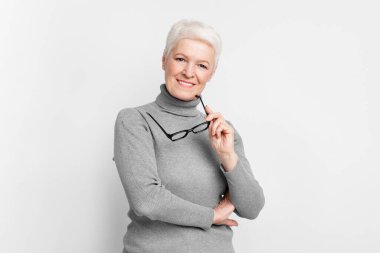 Gri arka planda gözlüklü yaşlı Avrupalı kadın, kameraya gülümsüyor. S3niorlife ve bilgelik temaları için mükemmel.