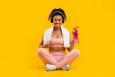 An african american lady in fitness attire sits cross-legged, smiling with a water bottle, isolated on yellow clipart