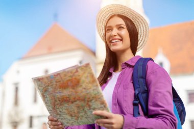 Hasır şapkalı bir turist kadın, açık gökyüzünün altındaki eski bir Avrupa şehir meydanında coşkulu bir beklentiyle bir harita okuyor.