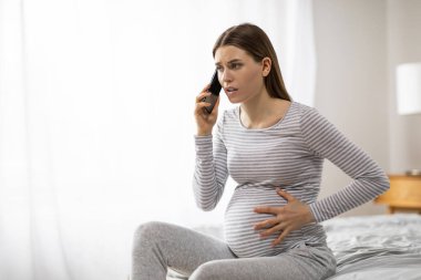 Çizgili elbiseli hamile bir kadın cep telefonuyla konuşurken, karnına dokunurken endişeli görünüyor.